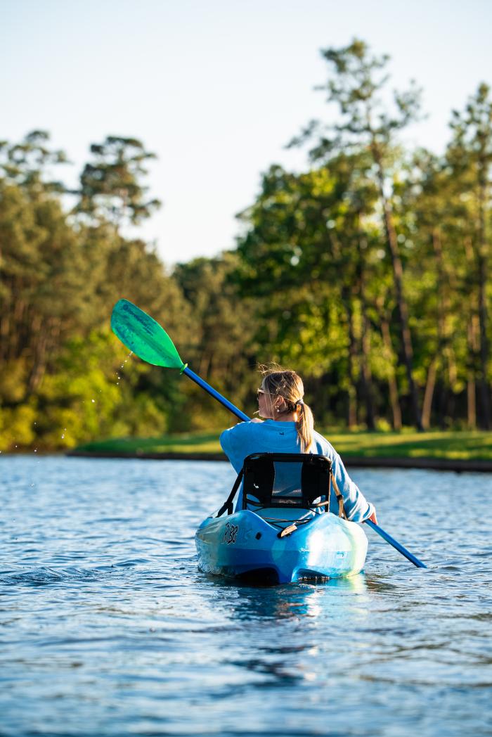 A young blonde woman (Brooklyn Newell) holds a blue and green double-sided paddle to row her bright blue kayak. She is facing away from the camera, paddling along Lake Woodlands toward a copes of trees.