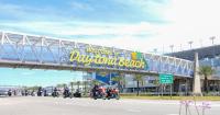 Motorcycle riders cruise under the Welcome to Daytona Beach sign at the Speedway under bright blue skies
