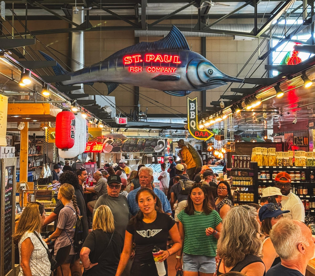 Visitors explore food vendors and shops inside the bustling Milwaukee Public Market beneath the large, illuminated St. Paul Fish Company sign shaped like a fish.