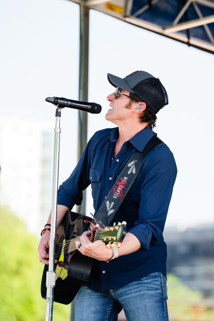 The lead singer of the Will Carter Band plays a black guitar on a stage at The Woodlands Waterway Arts Festival. He's wearing a navy button-down, blue jeans, and a dark ballcap. The guitar strap is embroidered with the band name and silver leaves.