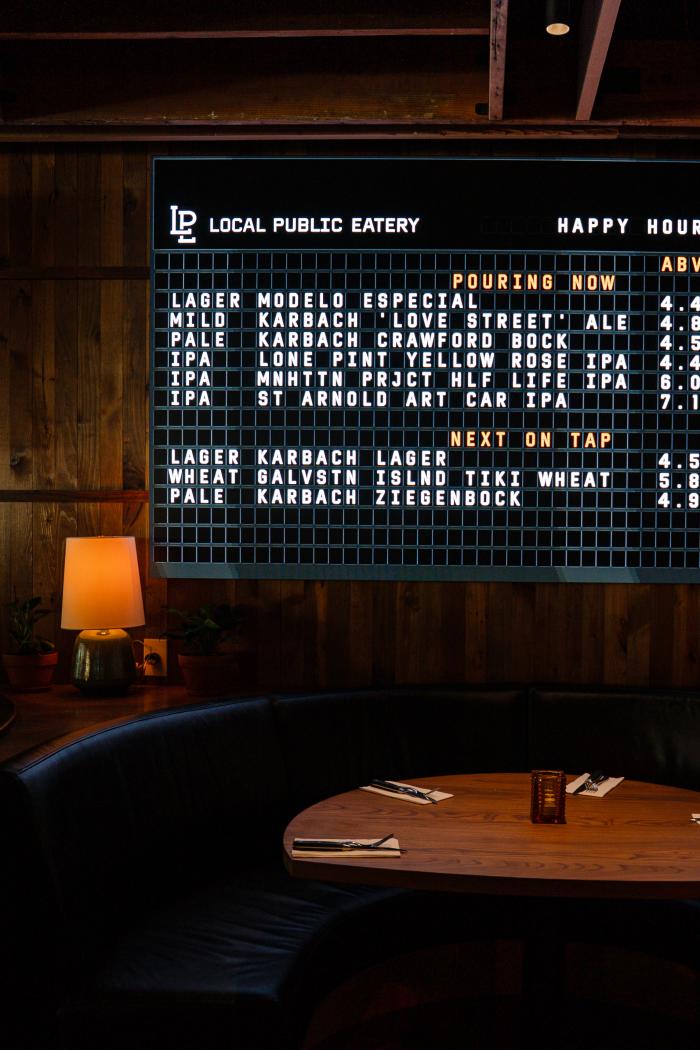Inside LOCAL Public Eatery is an electronic letter board featuring the bar's happy hour beer options. Below the board is a blue-padded booth with mood lighting.