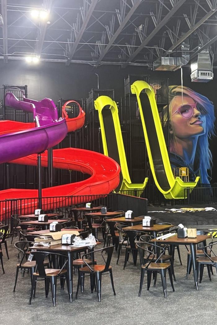 A phone photo of various slides at Slick City Action Park. On the left are two interlocking slides, one red and one purple. In the middle, two yellow "jump" slides. On the right, a mural of a young woman with pink glasses and blue highlights in her blonde hair.