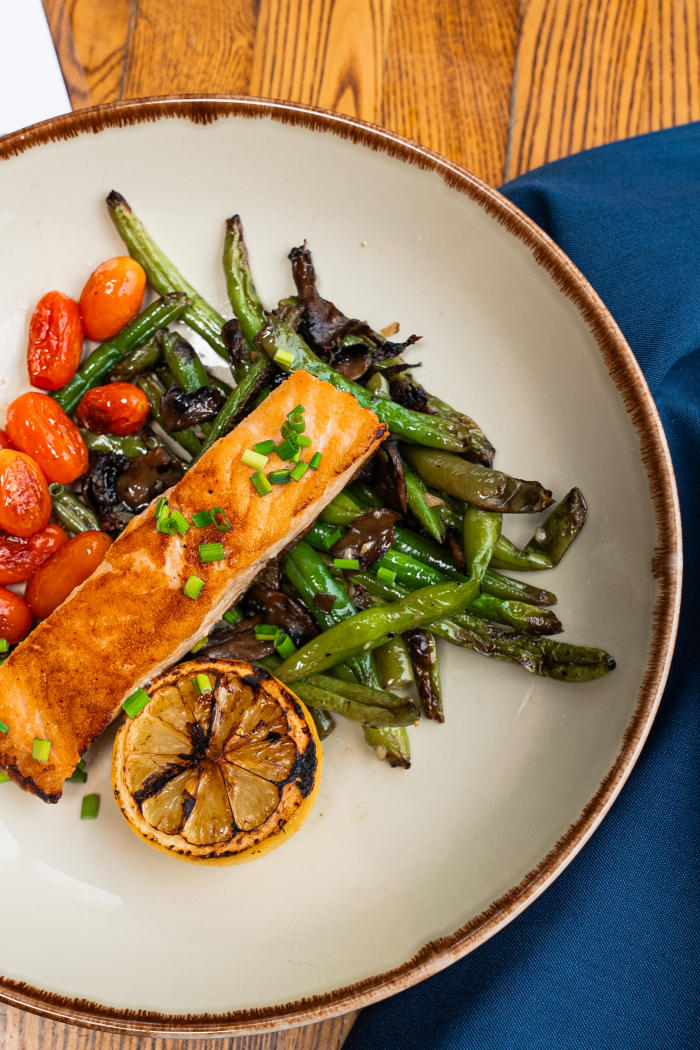 An overhead view of a white plate with grilled salmon garnished with roasted asparagus, tomatoes, and a lemon slice. The plate is resting on a blue cloth napkin.