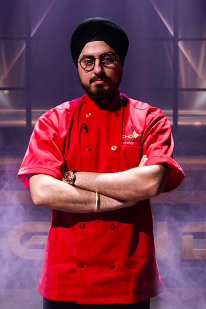 A 3/4 shot of Chef Jassi Bindra on a competitive TV cooking show. He's posing with his arms crossed, wearing a black turban and a bright red uniform with "CHOPPED" over the breast pocket. Purple lights and stage smoke fill the background.