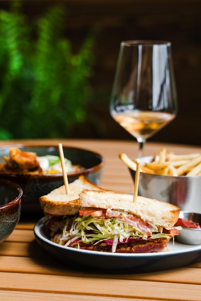 A food spread on the patio at LOCAL Public Eatery features a thick BLT and fries, a bowl of gourmet ramen, a bowl of fried chicken and rice, a beer in a plastic cup, and a small glass of white wine.
