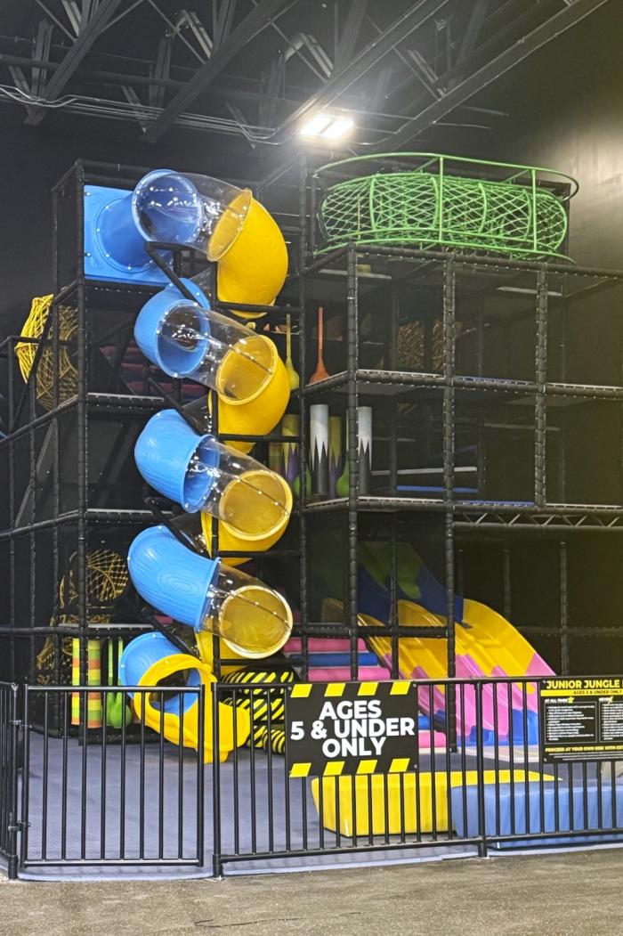 A phone photo of the Junior Jungle at Slick City Action Park. On the left is a twisty slide alternating in blue, yellow, and clear sections. Young children can access this slide by first climbing through a jungle gym.