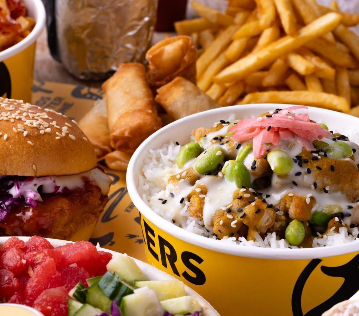 Large spread of food on a wooden plank table that includes a katsu curry bowl buddah bowl fries spring rolls and duck burger from Kwackers