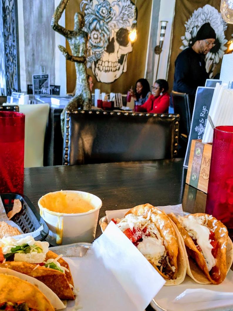 A table full of tacos, queso, red glasses, with funky art on the walls in the background at Agave and Rye in Covington, Ky.