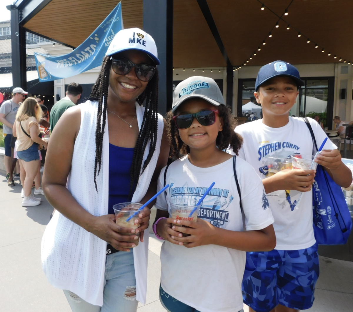 A smiling woman and two children enjoy frozen drinks on a sunny day. The woman wears sunglasses, a white vest, and a blue and white “I ❤️ MKE” hat. One child wears a Lakeshore Chinooks hat and sunglasses with a t-shirt that reads “I celebrated my birthday at Aquatics Department,” while the other wears a Stephen Curry shirt and a dark blue baseball cap. They are standing outside near a concession stand with other people in the background under a string of outdoor lights.