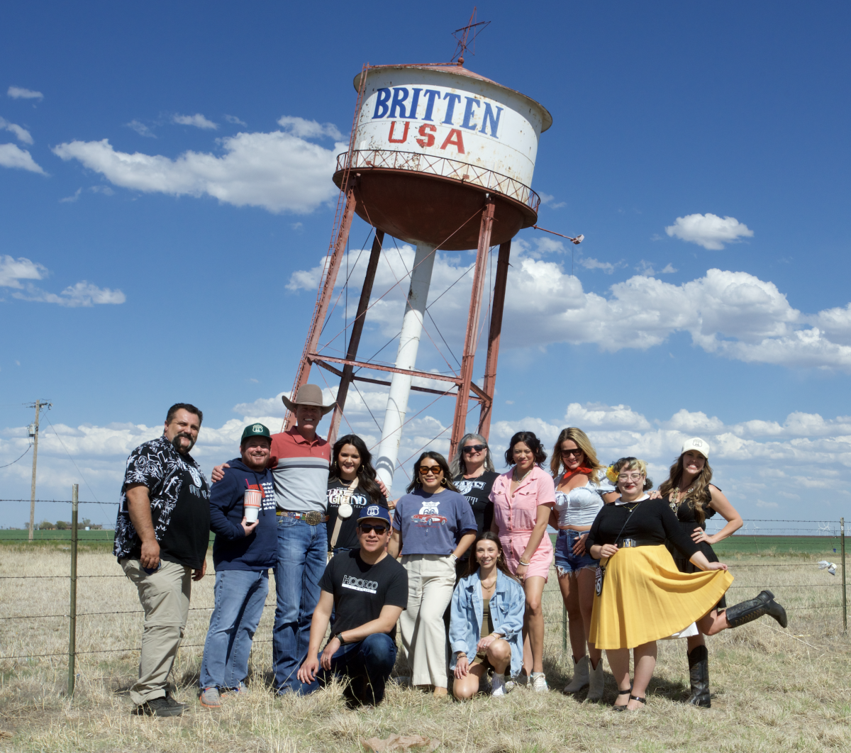 A group of people pose smiling in front of the iconic leaning Britten USA water tower, a quirky roadside attraction along Route 66 near Amarillo. Set against a wide blue Texas sky and open prairie, the slanted structure and cheerful group capture the fun, offbeat spirit of the Texas Panhandle.