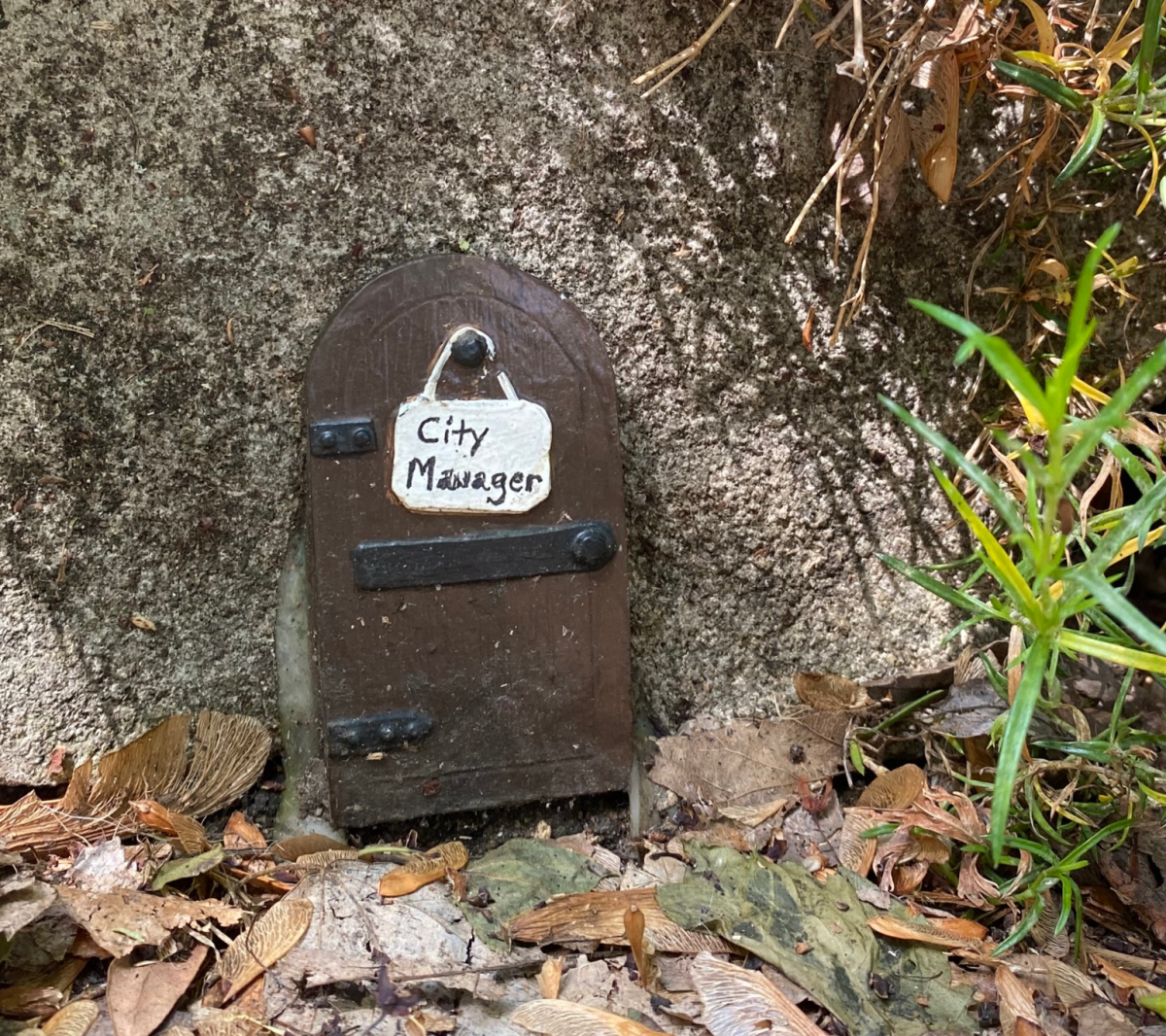 Image is of a small, brown fairy door that say's "City Manager" on it.