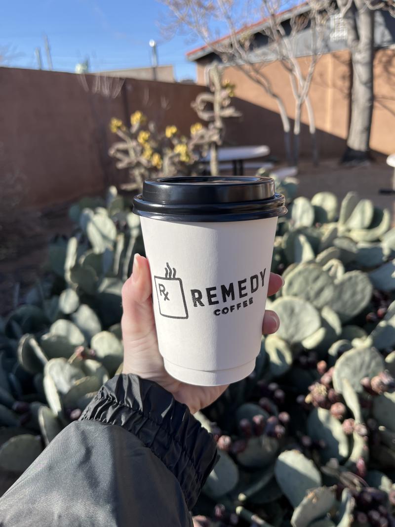 Someone holds a hot coffee cup from Remedy Coffee in front of a cactus.