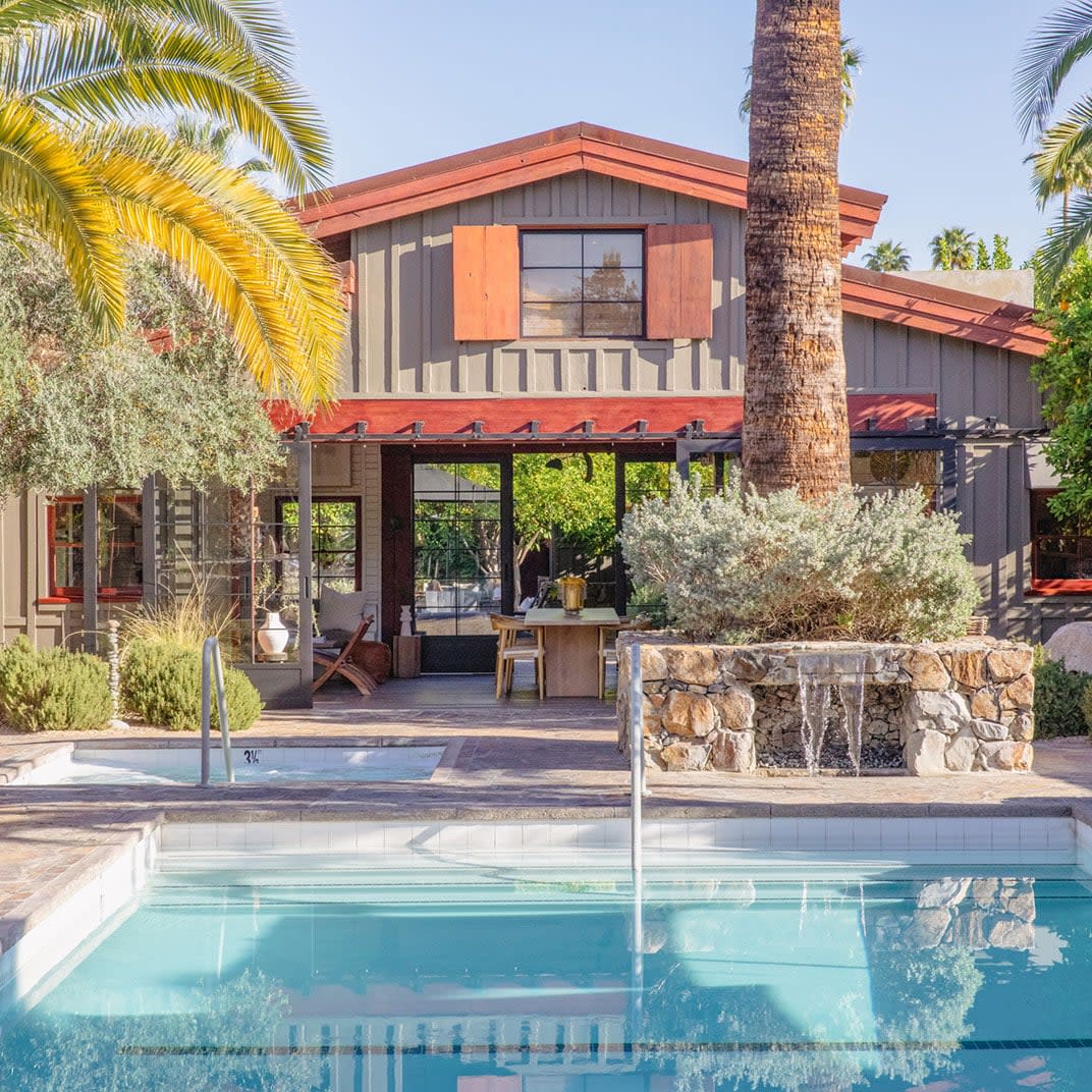 A two-story house with a mix of modern and rustic design elements, featuring wood paneling and large windows, set behind a swimming pool with a small waterfall feature, surrounded by lush palm trees under a clear blue sky.