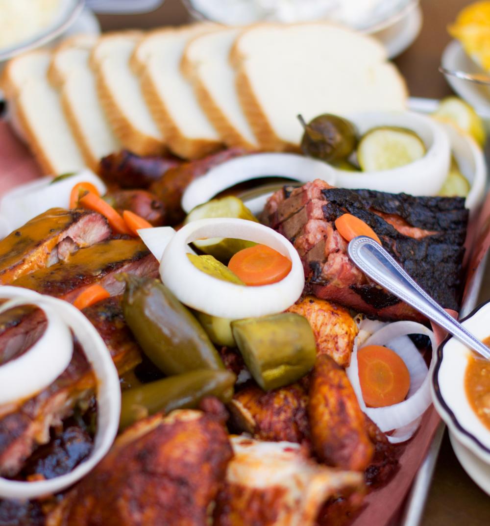 Metal platter of various meats and pickled vegetables surrounded by slices of white bread and bowls of sauces.