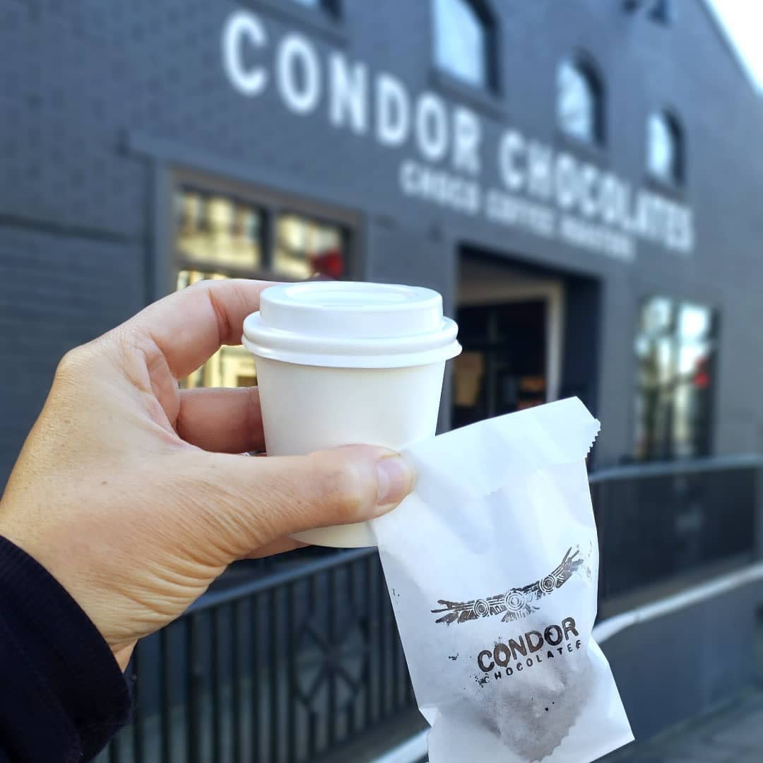 A hand holds a to-go cup and small Condor Chocolates bag in front of Condor's new downtown store front.