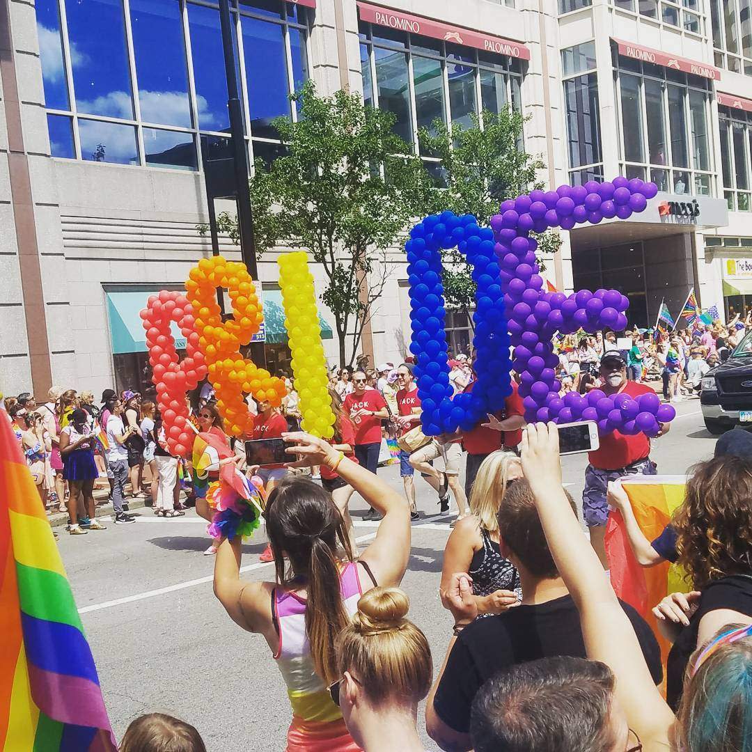 Image is of the Cincinnati Pride Parade with a group carrying the letters P.R.I.D.E made of ballons as they walk down the street.