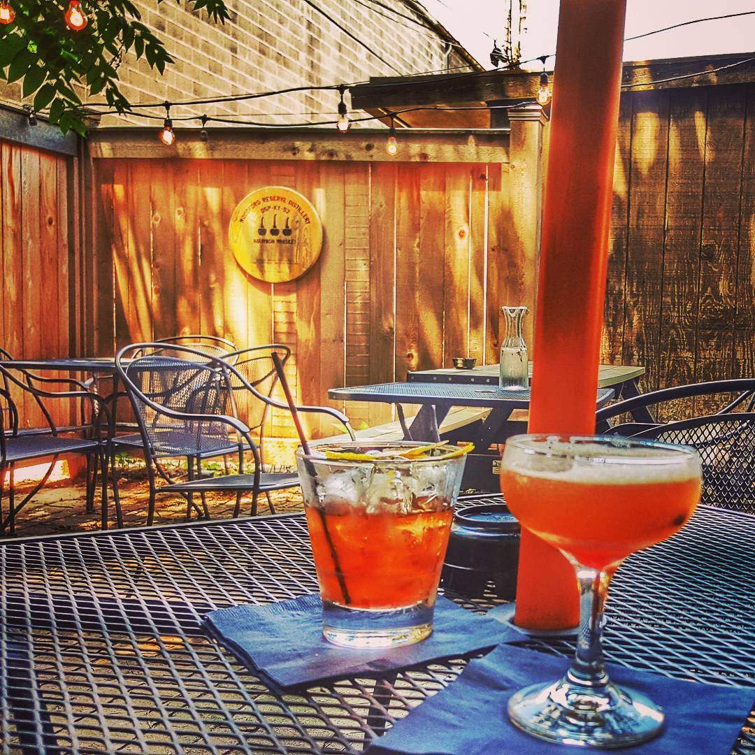A wooden patio with sunshine and two peach-colored cocktails on an iron table