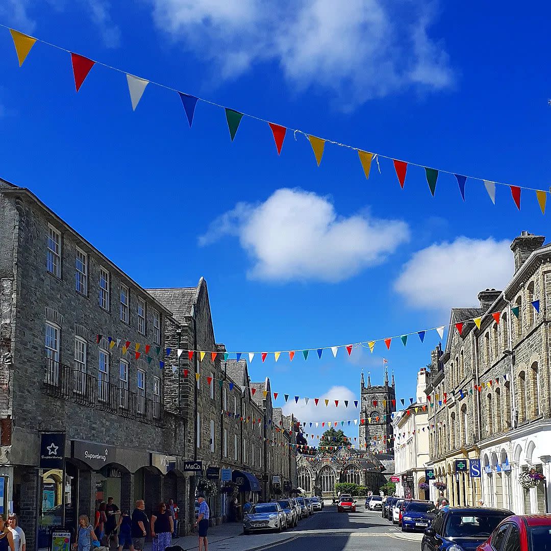 A vibrant street scene with colorful pennant flags strung between gray stone buildings under a bright blue sky. People and parked cars line the street.