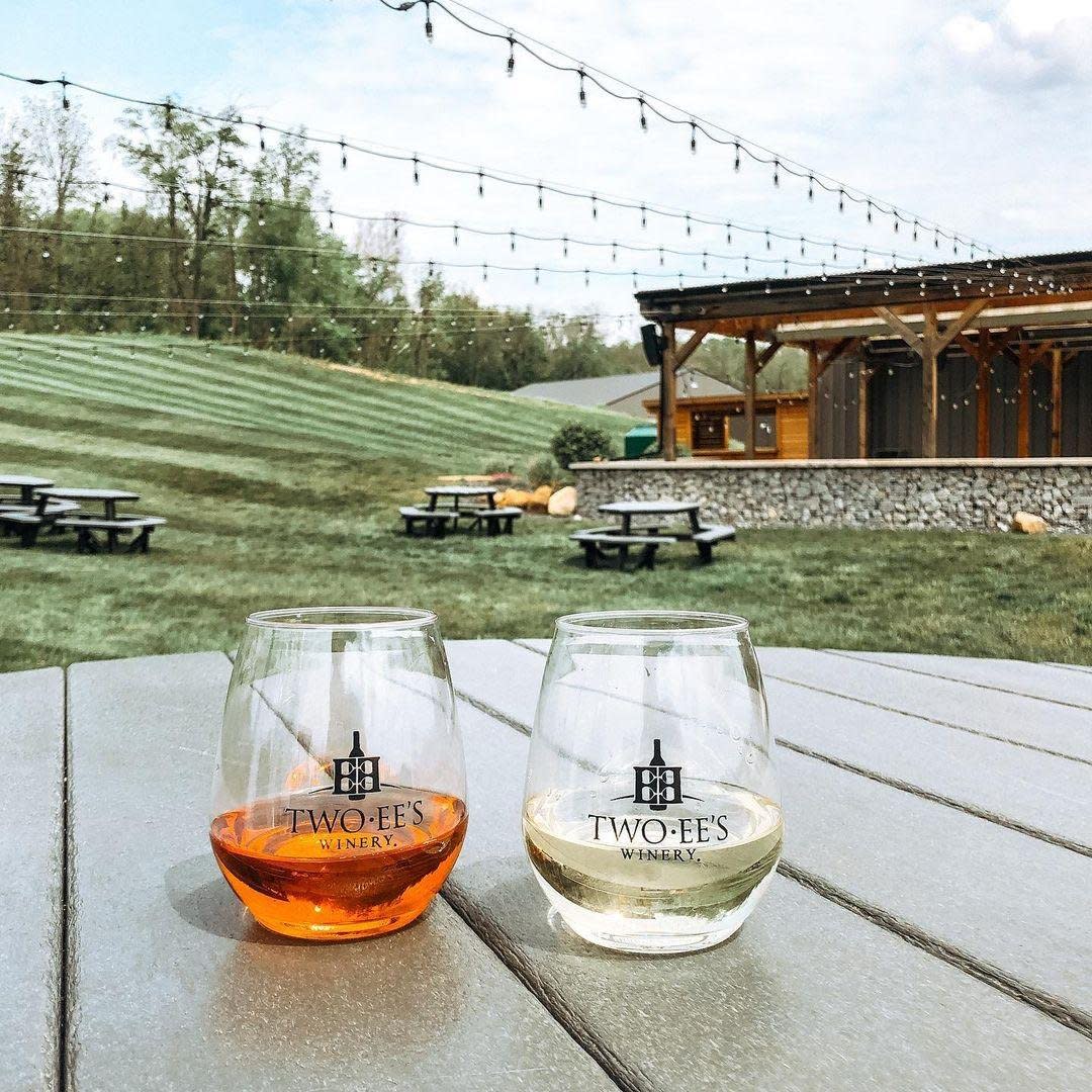 two stemless wine glasses on a table outside at two ees winery. one glass has rose wine and the other has white wine. You can see string lights, grass, and the winery building in the background