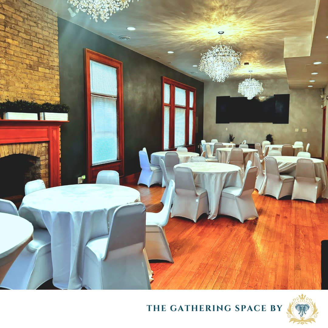 A banquet-style event room with hardwood floors, round tables with white linens, white chair covers, crystal chandeliers, tall windows, a brick fireplace, and a large wall-mounted TV.