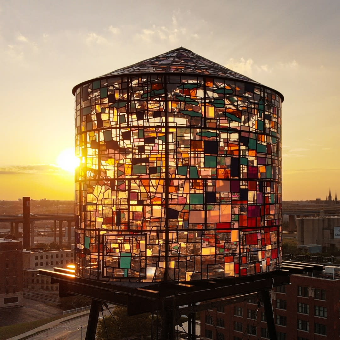 A colorful water tower sculpture made of multicolored glass panels glows in the light of the setting sun. The translucent panels in shades of red, orange, blue, green, and purple create a stained-glass effect as sunlight filters through. The sculpture sits atop a rooftop in Milwaukee’s Historic Third Ward, overlooking city buildings and bridges in the background. The warm golden sky enhances the luminous effect of the artwork.