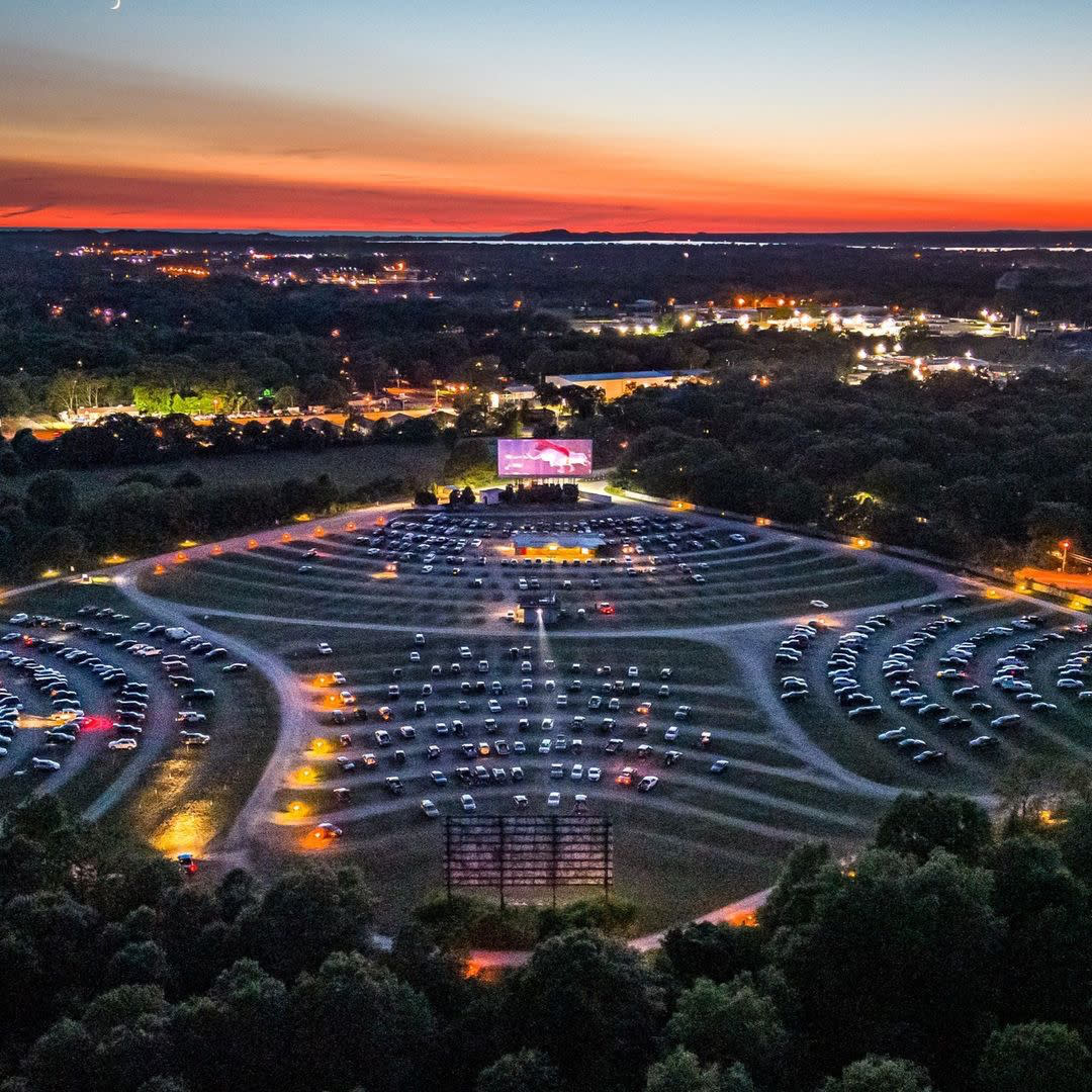birds eye viewof Getty drive in theater at sunset in muskegon michigan shows cars parked at 4 screens surrounded by trees, city lights in distance and sunset on horizon