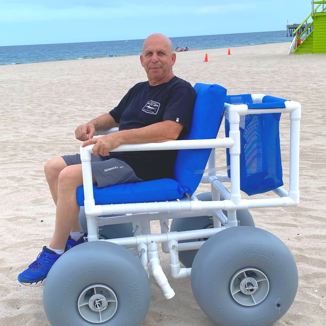 A man using a beach wheelchair. The chair is made of PVC and has four grey balloon-like rubber wheels to enable the chair to roll across the sand