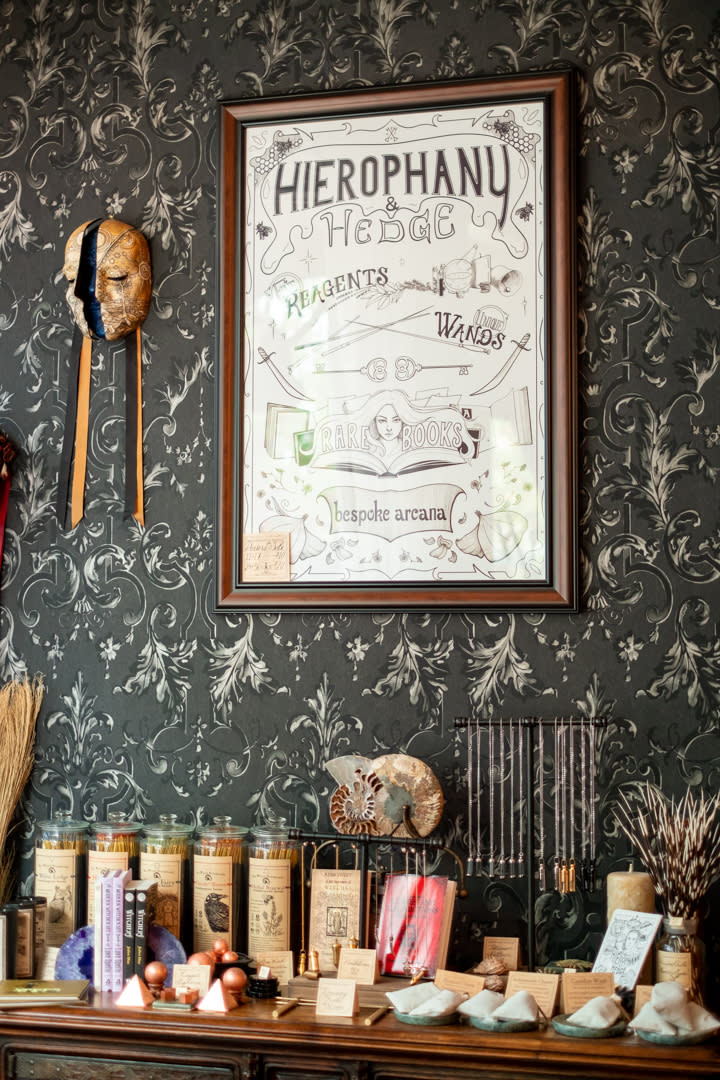 A poster that reads Hierophany and Hedge, reagents, wands, bespoke arcana, hanging on a dark wallpapered wall