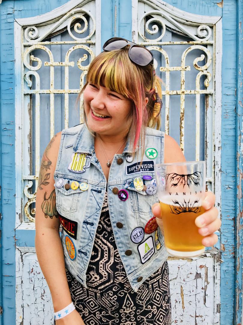 A woman in a denim sleeveless shirt stands in front of an iron gate painted white holding a beer glass that reads "Vanish"