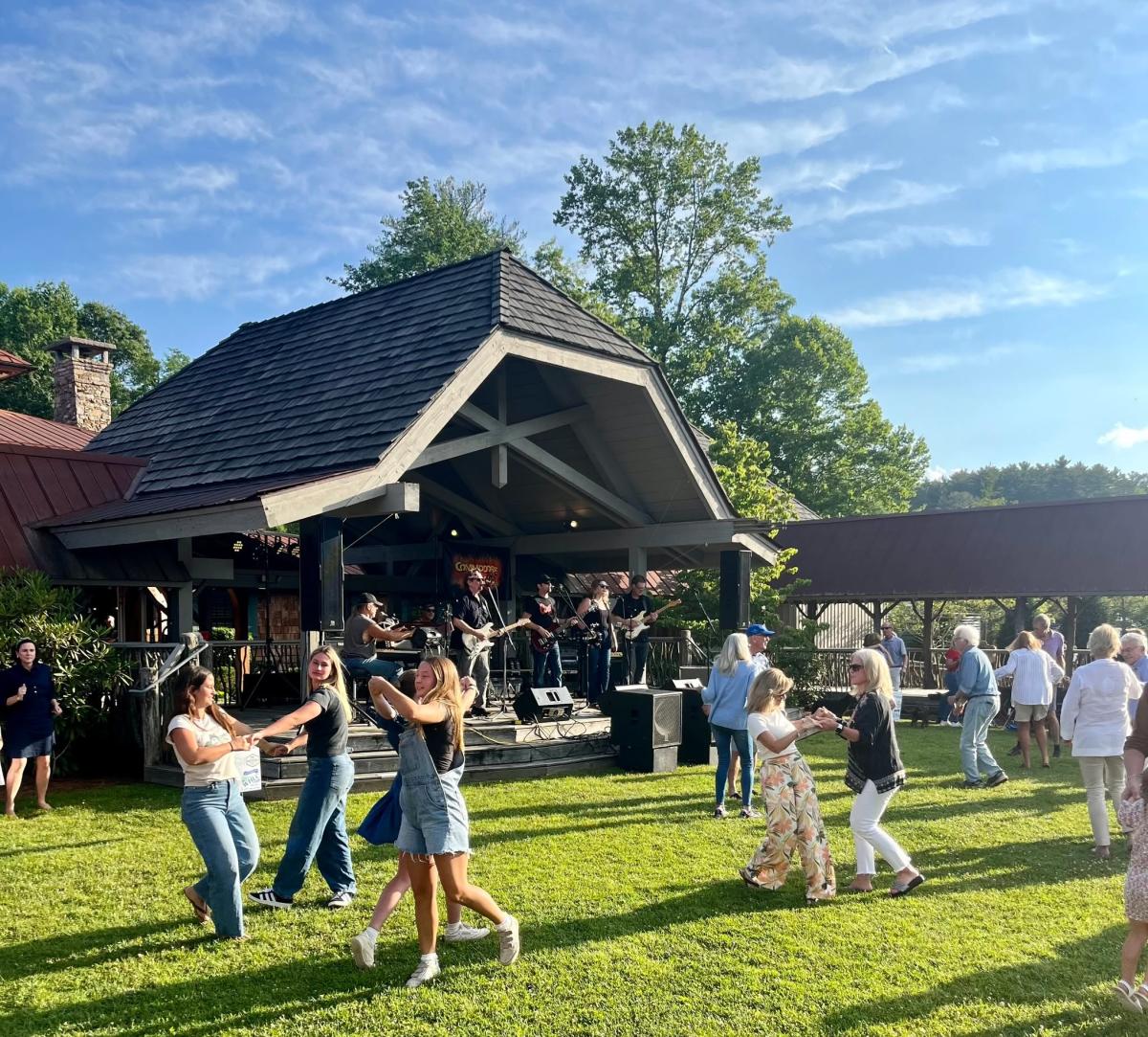 A band plays live music on a rustic wooden stage under a pavilion. Multiple groups of people, including teenagers on the left wearing denim and adults on the right wearing casual attire, dance enthusiastically on a bright green grassy lawn on a sunny day. Trees frame the background.