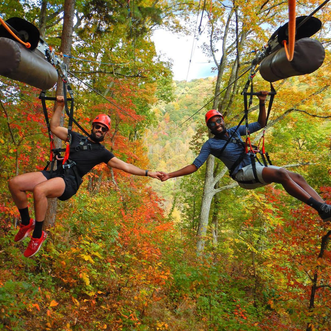 Two men holding hands ziplining through forest with fall-colored leaves at Navitat