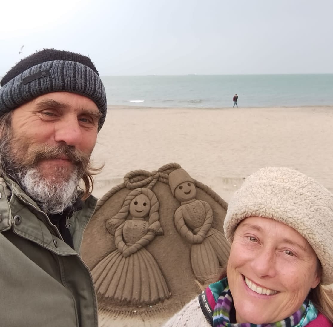 Selfie of a man and a women in beanies smiling at the camera while on the beach. Between them in the sand is a sculpture showing a man and a woman in a frame with bow detailing.