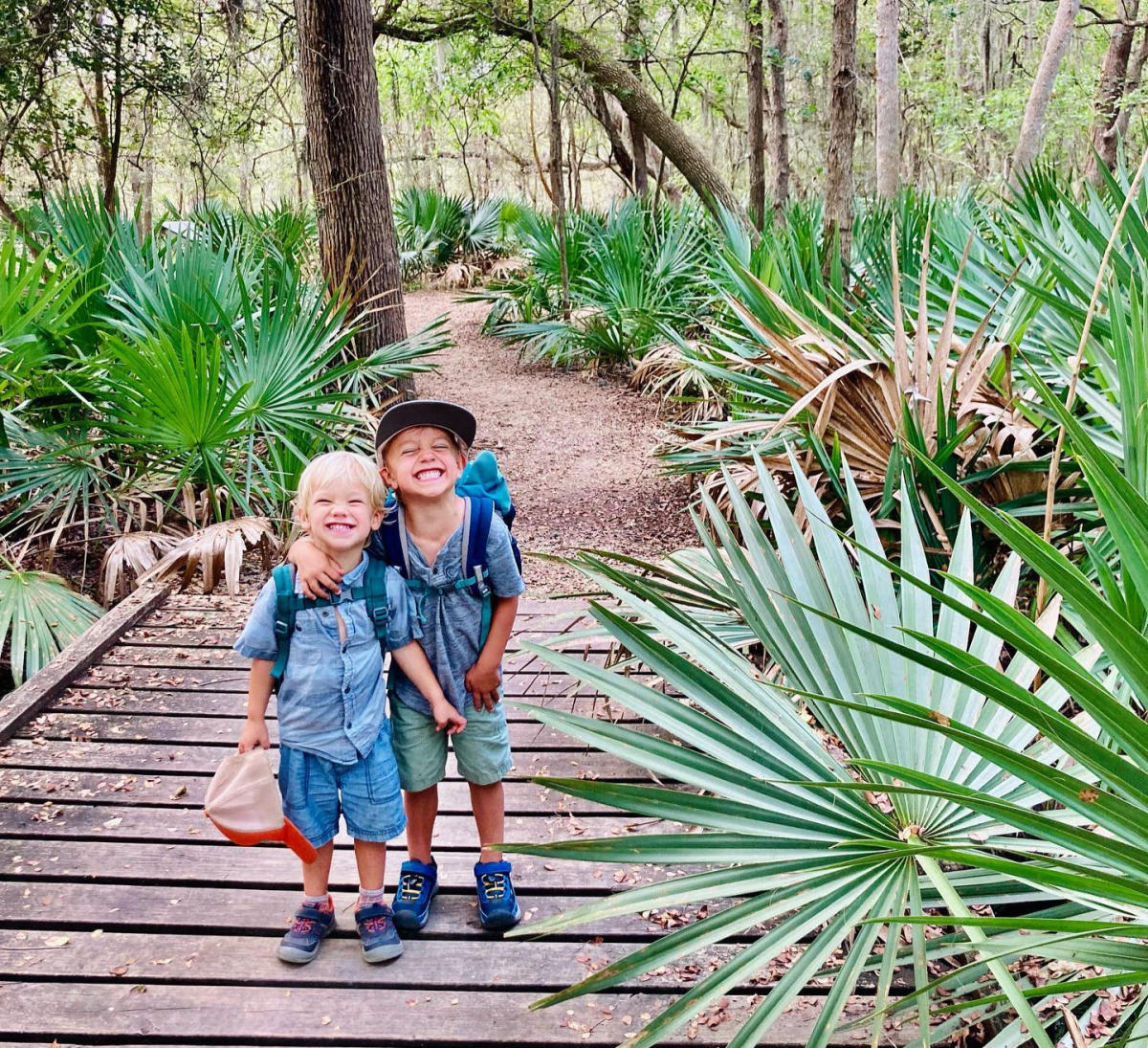 Two little boys in shorts and hiking shoes smiling at the camera surrounded by Palmetto Palms at Palmetto State Park.