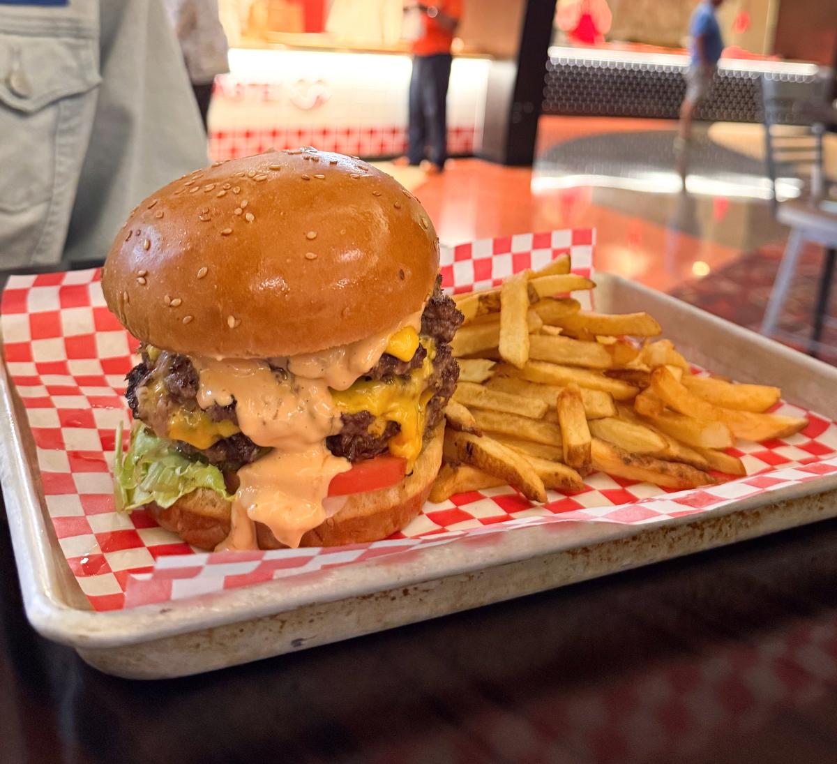 A tray lined with red and white checkered paper holds a cheeseburger stacked with two beef patties, melted cheese, lettuce, tomato, and sauce dripping from the bun, served with a side of golden French fries.