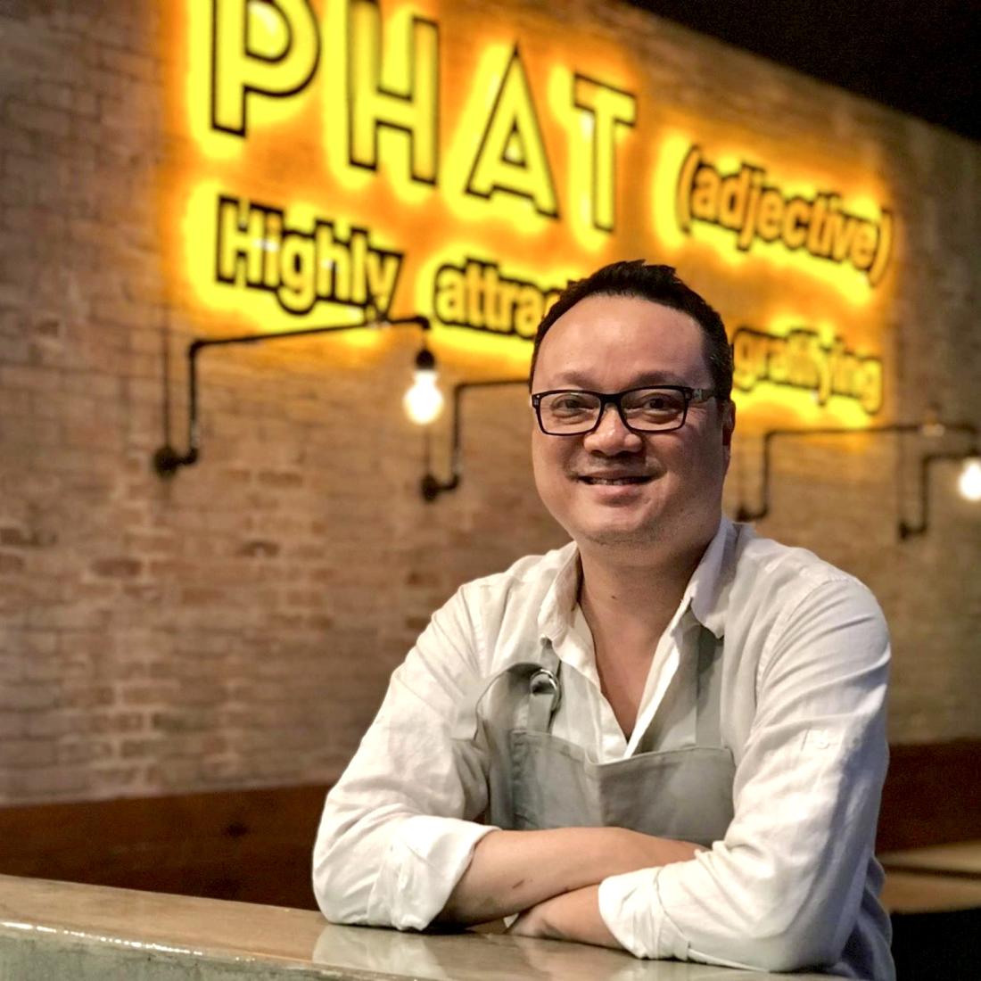 Alex Au-Yeung smiles at the camera. He has short black hair, black-rimmed glasses, and is wearing a white long-sleeved shirt with a light gray apron. He has his arms folded casually against one of the tables in his restaurant. Behind him is a yellow neon sign that says, "PHAT (adjective) Highly attractive or gratifying."