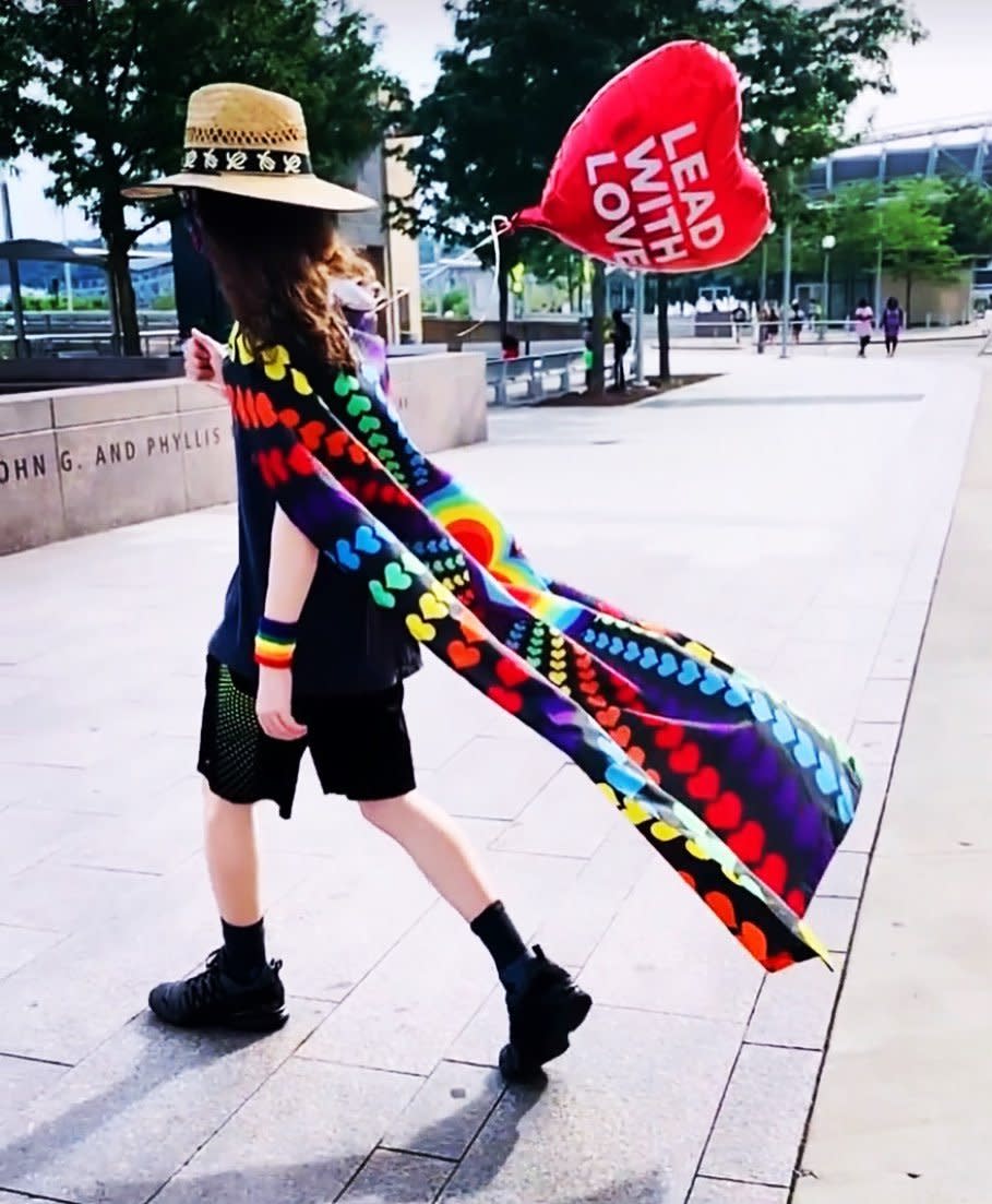 A young person with a straw hat in black t-shirt and shorts, wearing a rainbow cape and carrying a red heart-shaped ballon that says, "Lead with Love."