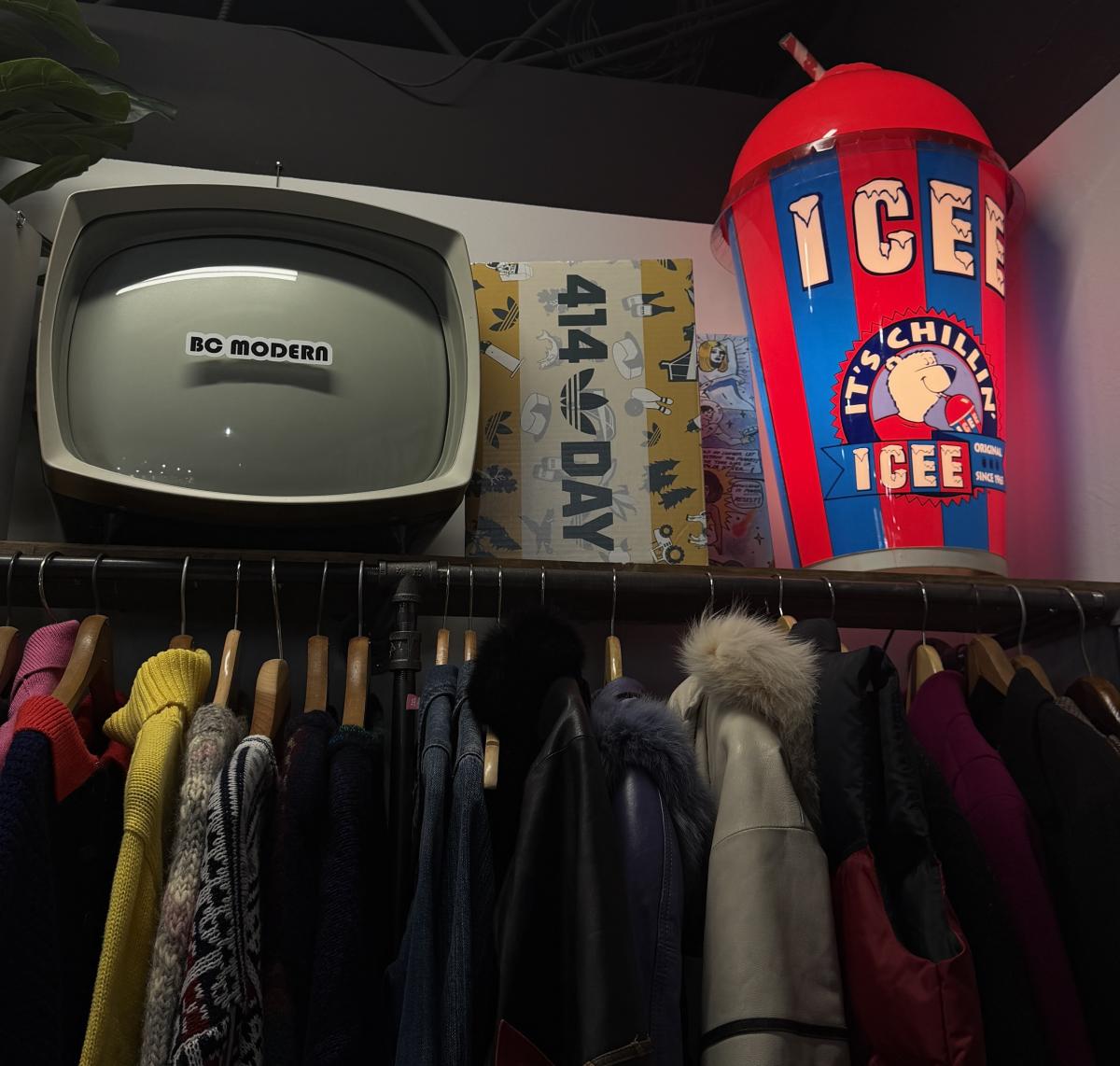Vintage clothing rack with coats and sweaters beneath a shelf displaying a retro CRT television labeled “BC Modern,” a “414 Day” poster, and a large colorful ICEE cup sign.