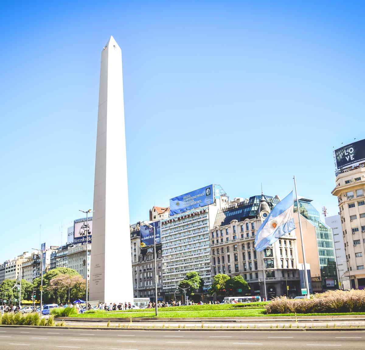 The Obelisk of Buenos Aires, a tall white monument, stands tall in the center of a busy city street. Buildings line the sides of the street, and an Argentine flag is visible flying from one of them.