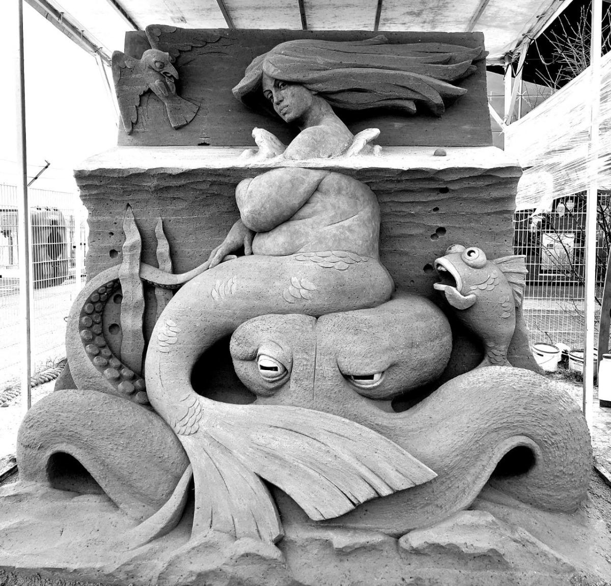 Black and white image of a sand sculpture that depicts a mermaid half in and half out of the water. She has rolls and is sitting on a disgruntled looking octopus. Other sealife is looking at her, shocked.