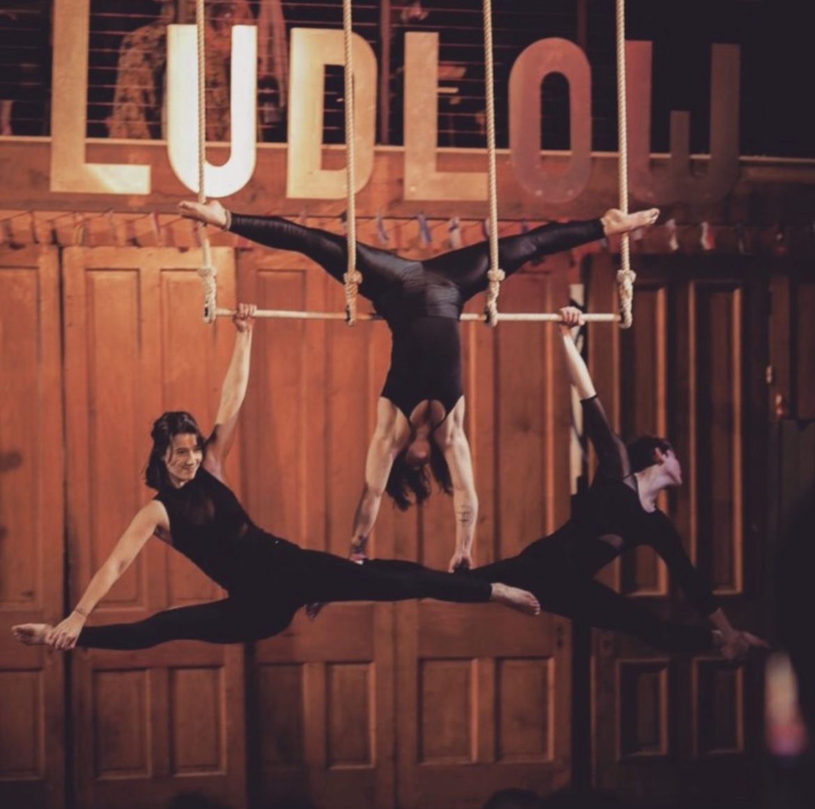 Image is of 3 trapeze artists in motion with the sign "Ludlow" behind them.