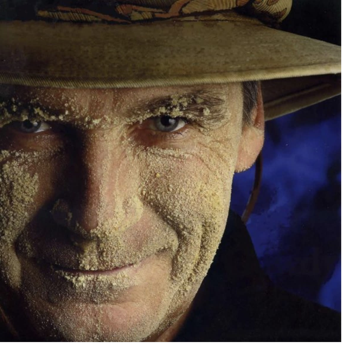 Close up headshot of a man smiling at the camera. He is wearing a hat and his face is covered in sand.