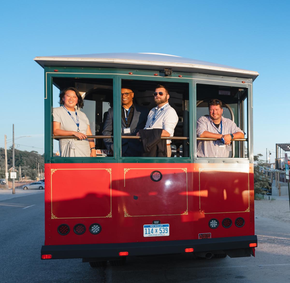 a woman and 3 men stand at rear of red and green trolley. all are smiling