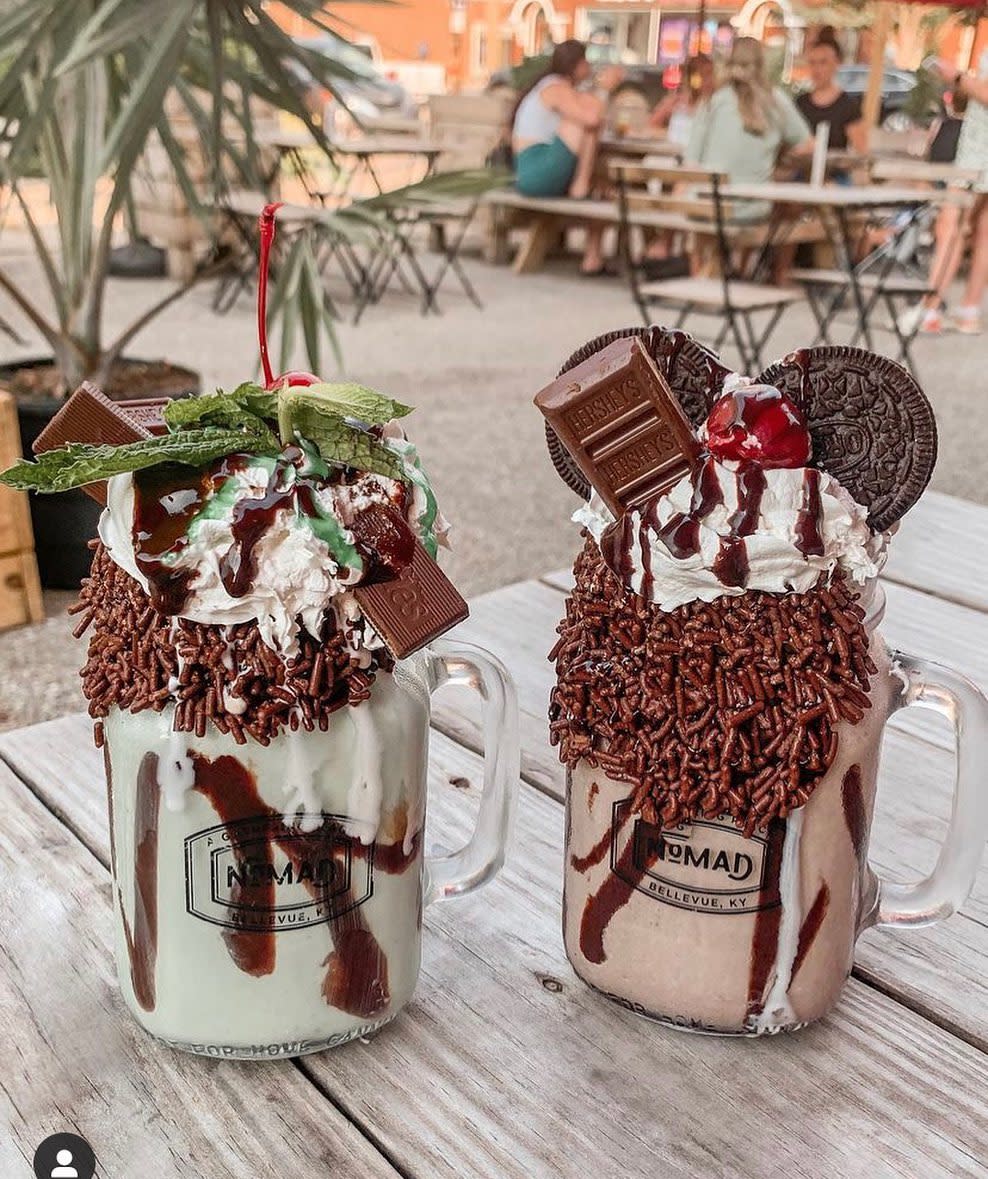 Two milkshakes, one mint chocolate, one chocolate, heaped with whipped cream and chocolate candies on a wood table outside at Nomad cafe in Bellevue, Ky.