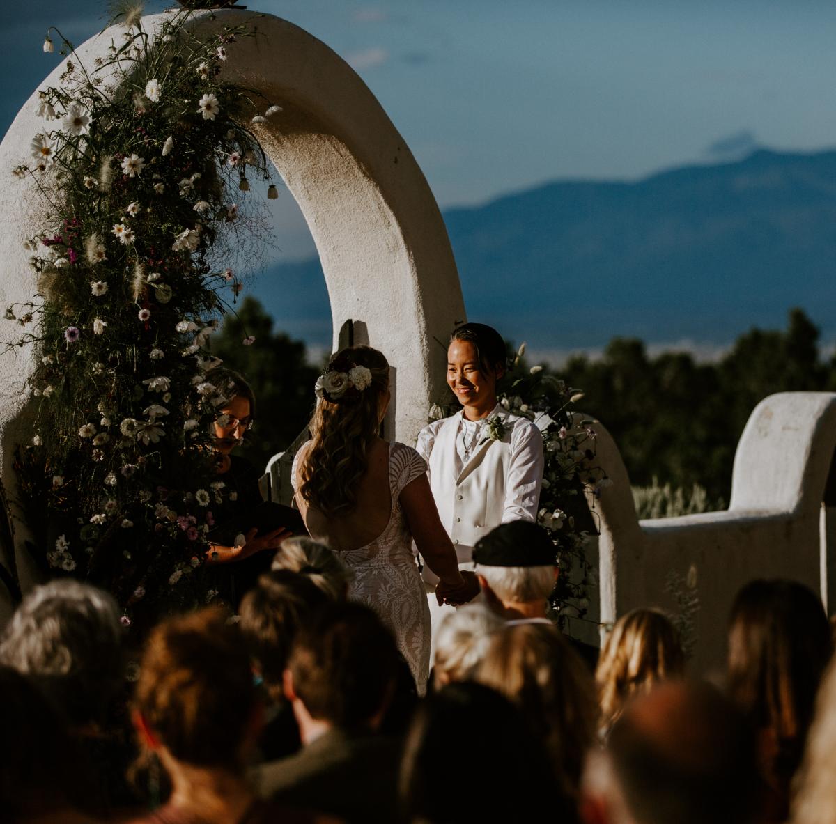 A couple exchanges vows at an outdoor wedding with mountains in the background. The scene is festive with guests seated and flowers adorning an arch.