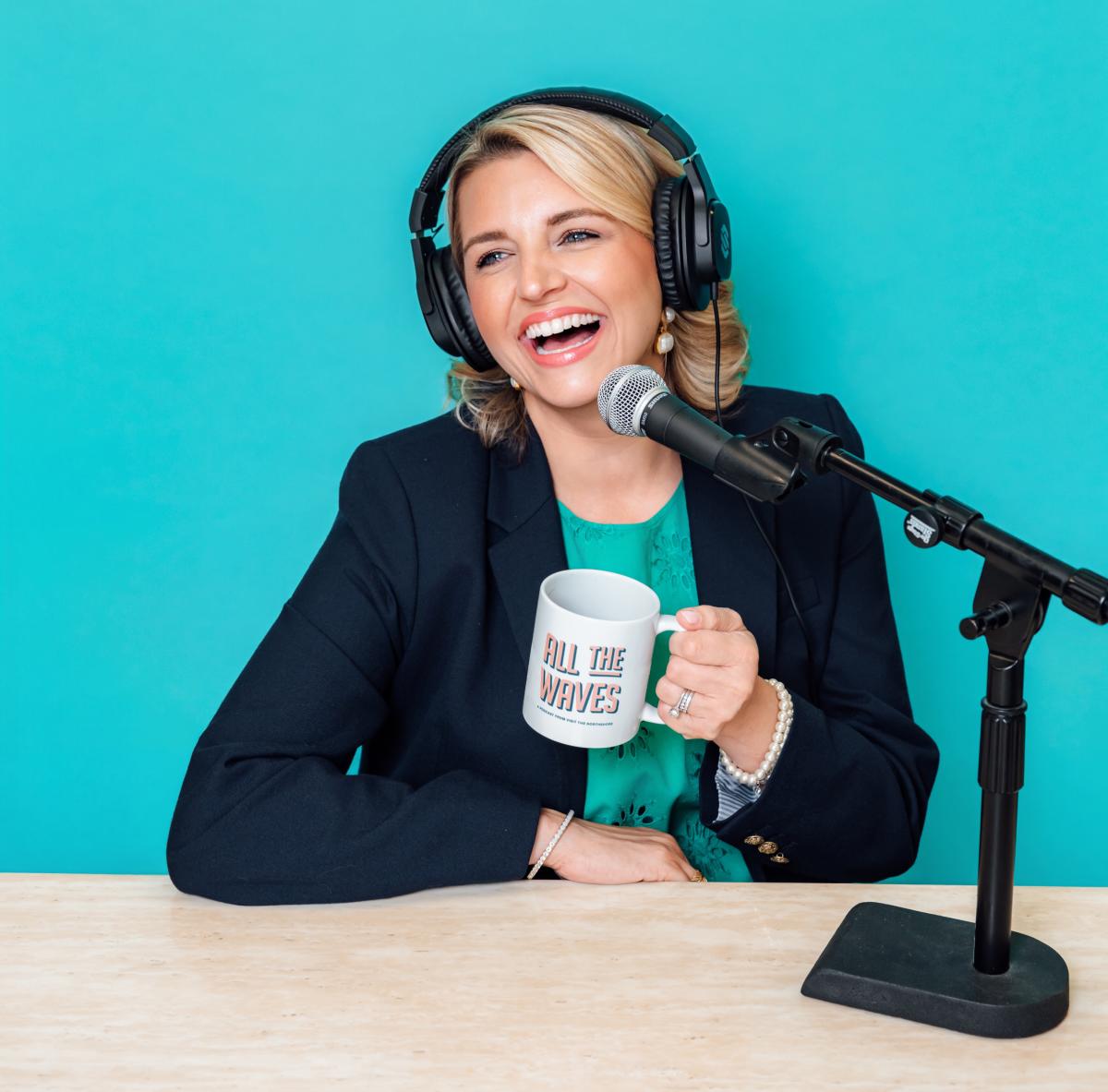 All The Waves co-host Katie Guasco laughs while recording an episode in the podcast studio. She is holding a logo mug.
