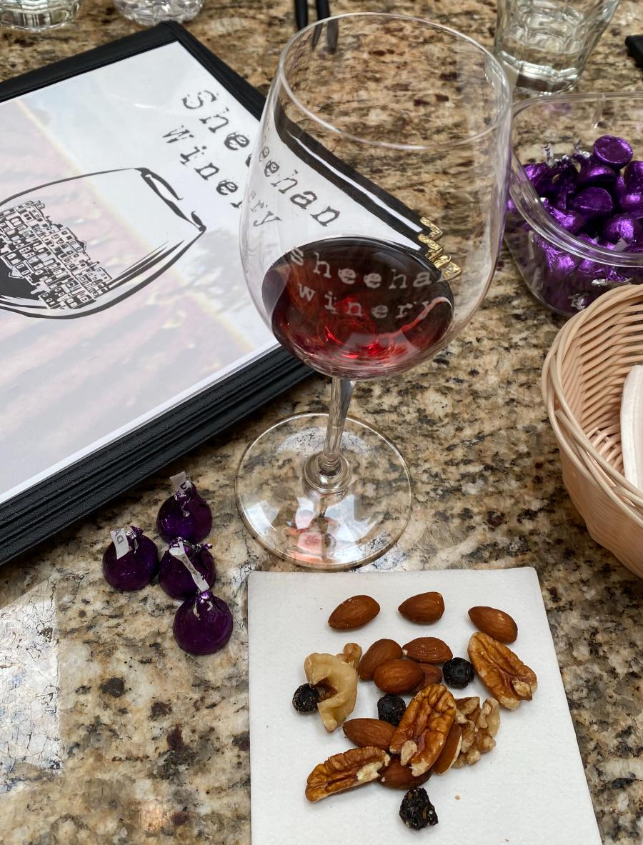 A Sheehan Winery menu, a glass of wine, some chocolate and some nuts sit on a table.