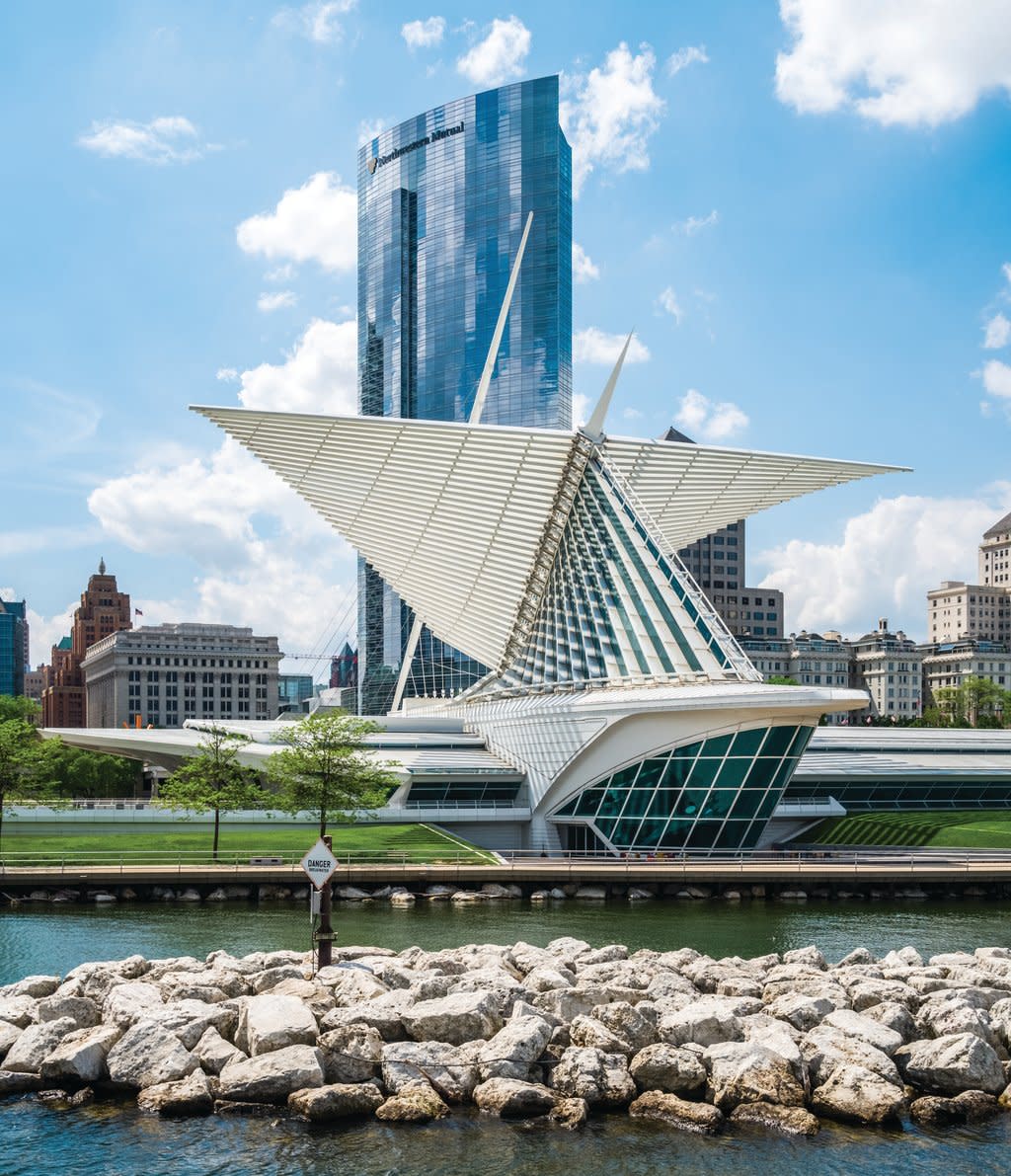 A daytime view of the Milwaukee Art Museum with its white, wing-like brise soleil extended against a blue sky. The sleek glass-and-steel structure sits along the lakefront, with a rocky shoreline in the foreground and tall downtown buildings rising behind it.