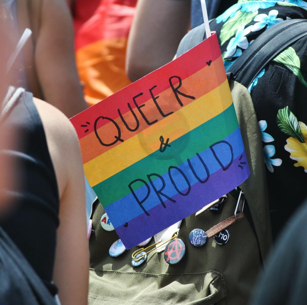 handheld Rainbow flag sticking out of a backpack in a crowd with "Queer & Proud" written on it