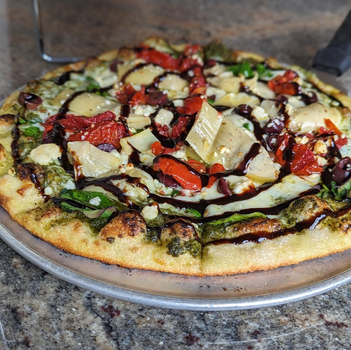 The image is of a Mediterranean pizza featuring a pesto sauce, blend of Italian cheeses, tomatoes and a balsamic glaze.