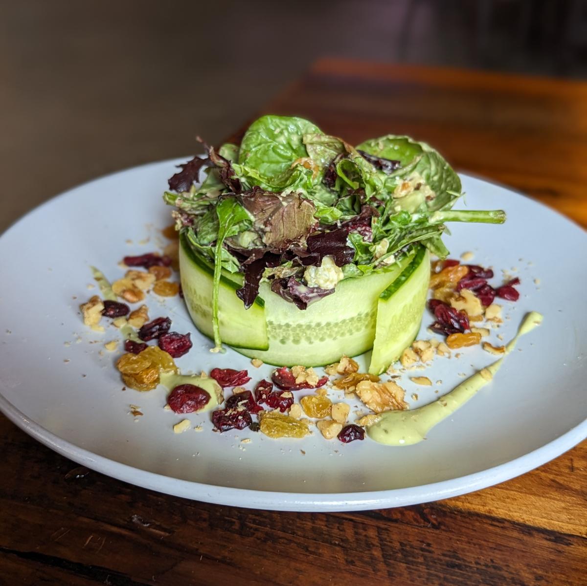 Image is of Shiners Green Machine salad which is a small salad that is wrapped together by a cucumber with raisins and dressing surrounding it.
